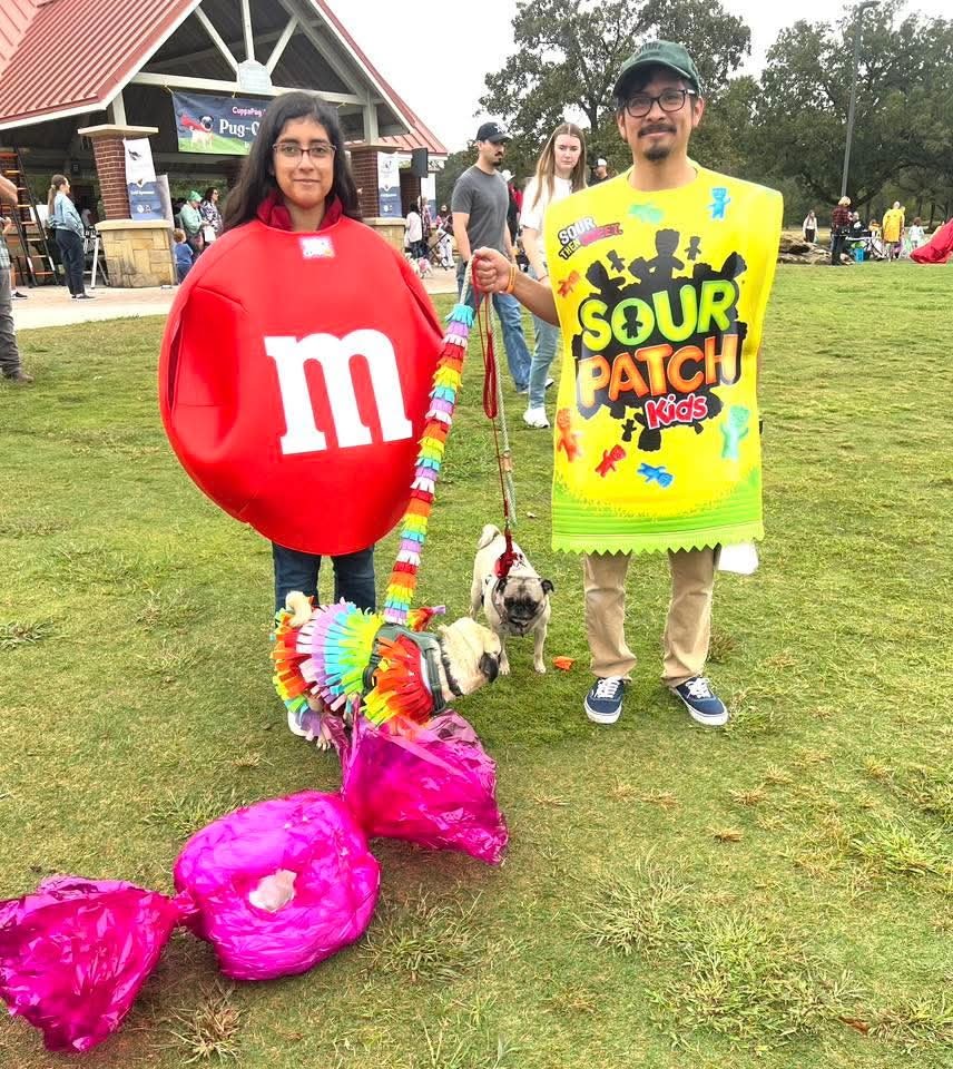 Two people dressed in costumes at an outdoor event. The woman is in a red M&M costume, and the man is dressed as Sour Patch Kids candy. They are holding leashes attached to a bulldog and a small dog, and there are pink balloon decorations on the ground.