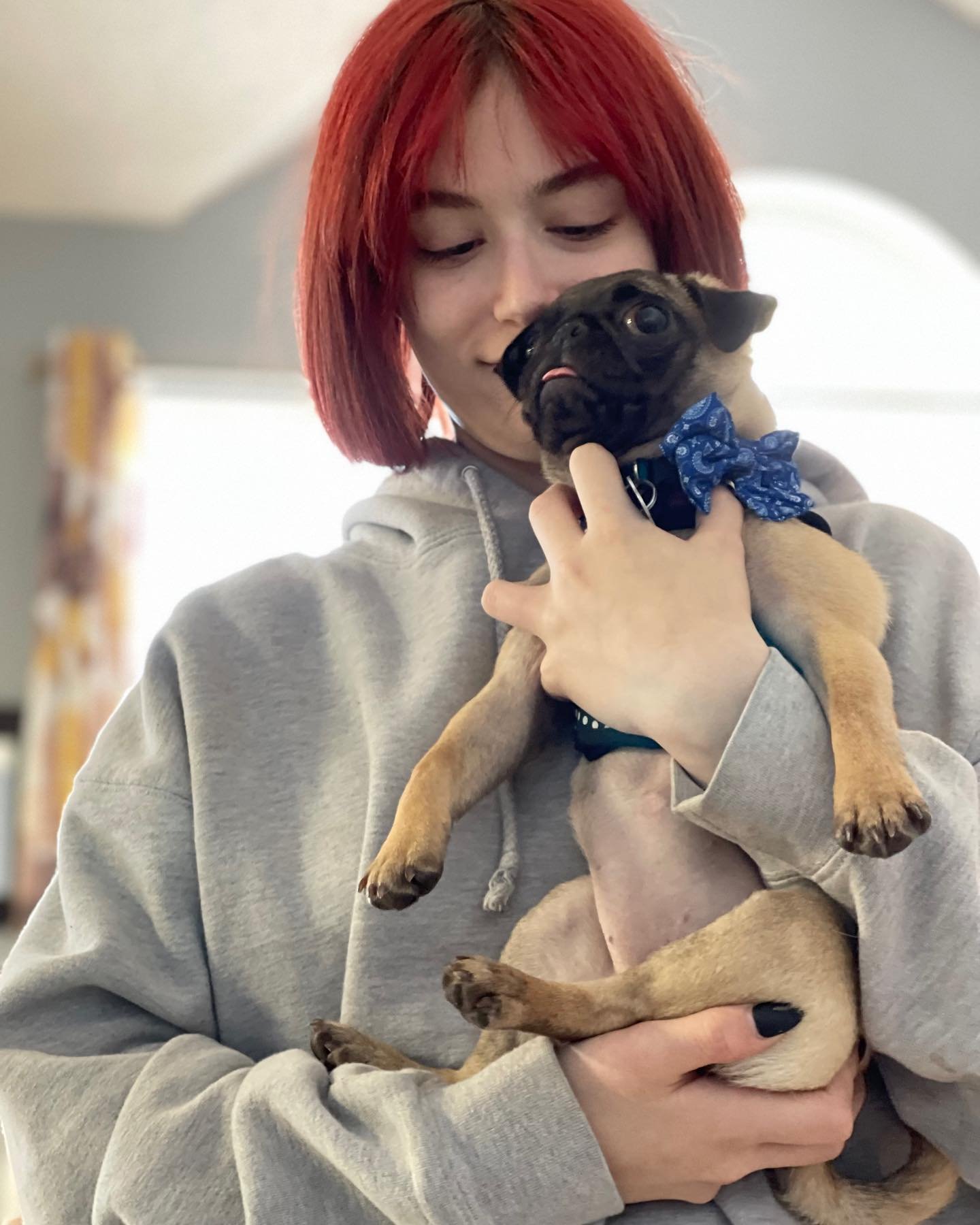 A woman with red hair holding a small tan and black French Bulldog puppy wearing a blue bow tie.