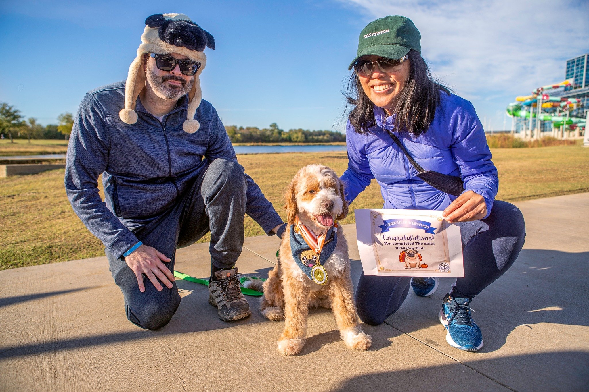 Two people kneeling on a sidewalk outdoors with a young dog wearing a medal and bandana, holding a certificate and smiling, in a park with waterpark slides in the background.