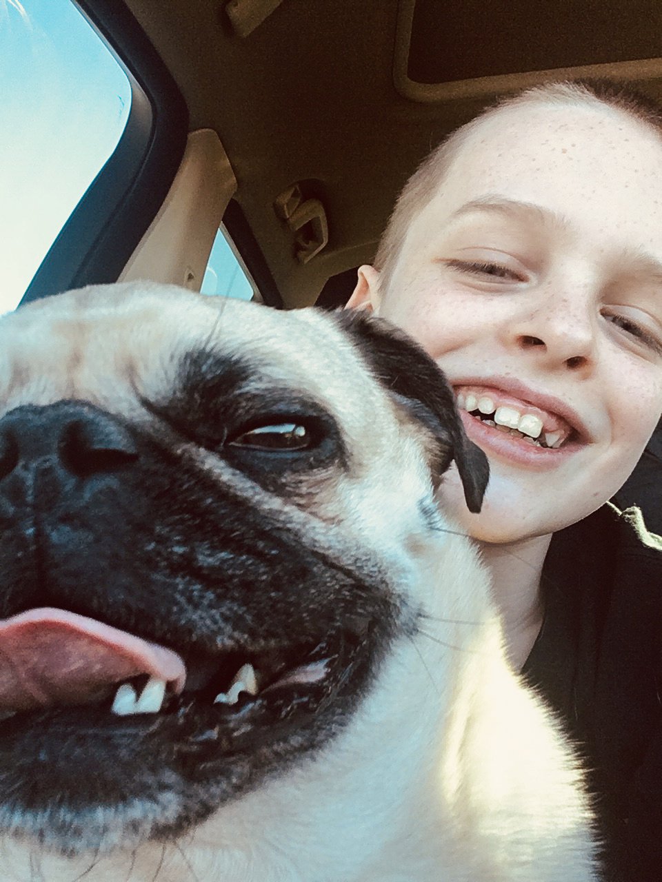 A boy with short light hair and freckles smiling with a dog, both sitting inside a vehicle. The boy has a missing tooth and is showing his teeth. The dog is a pug with a beige and black coat, tongue slightly out, and eyes partly closed.