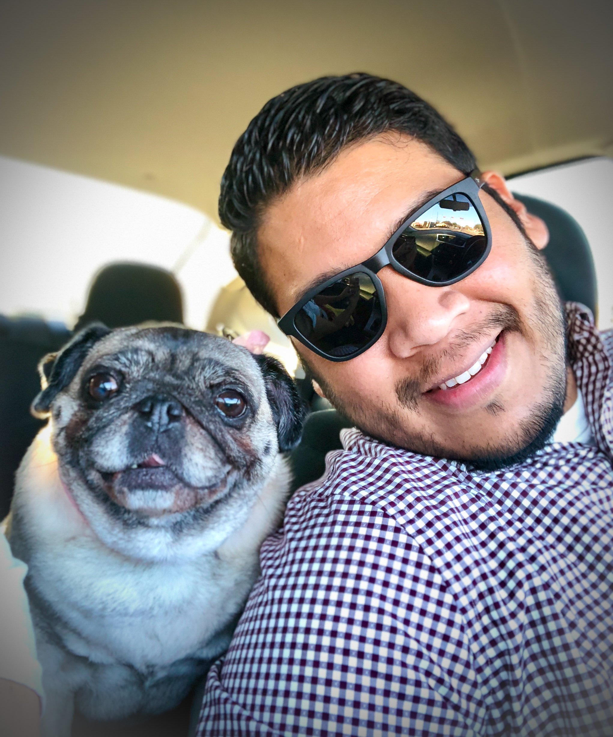 Man with sunglasses smiling, sitting beside a pug dog in a car.