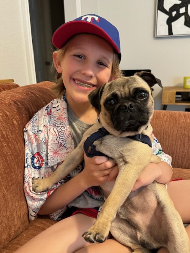A smiling young girl in a baseball cap holding a pug puppy on a brown couch in a living room.