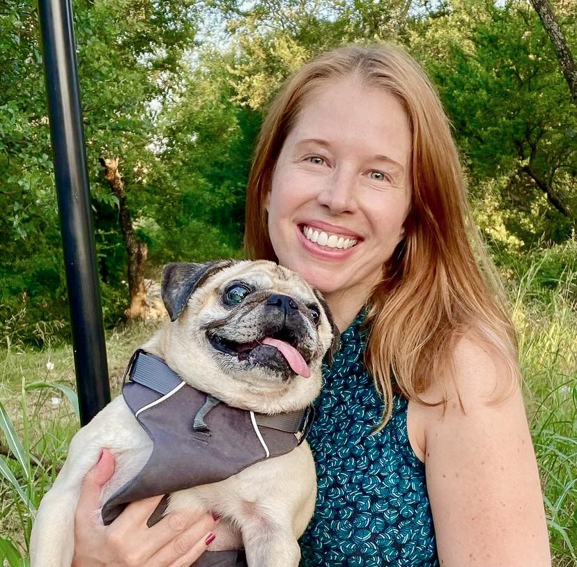 A smiling woman with long red hair holding a happy pug dog outdoors surrounded by green trees and grass.