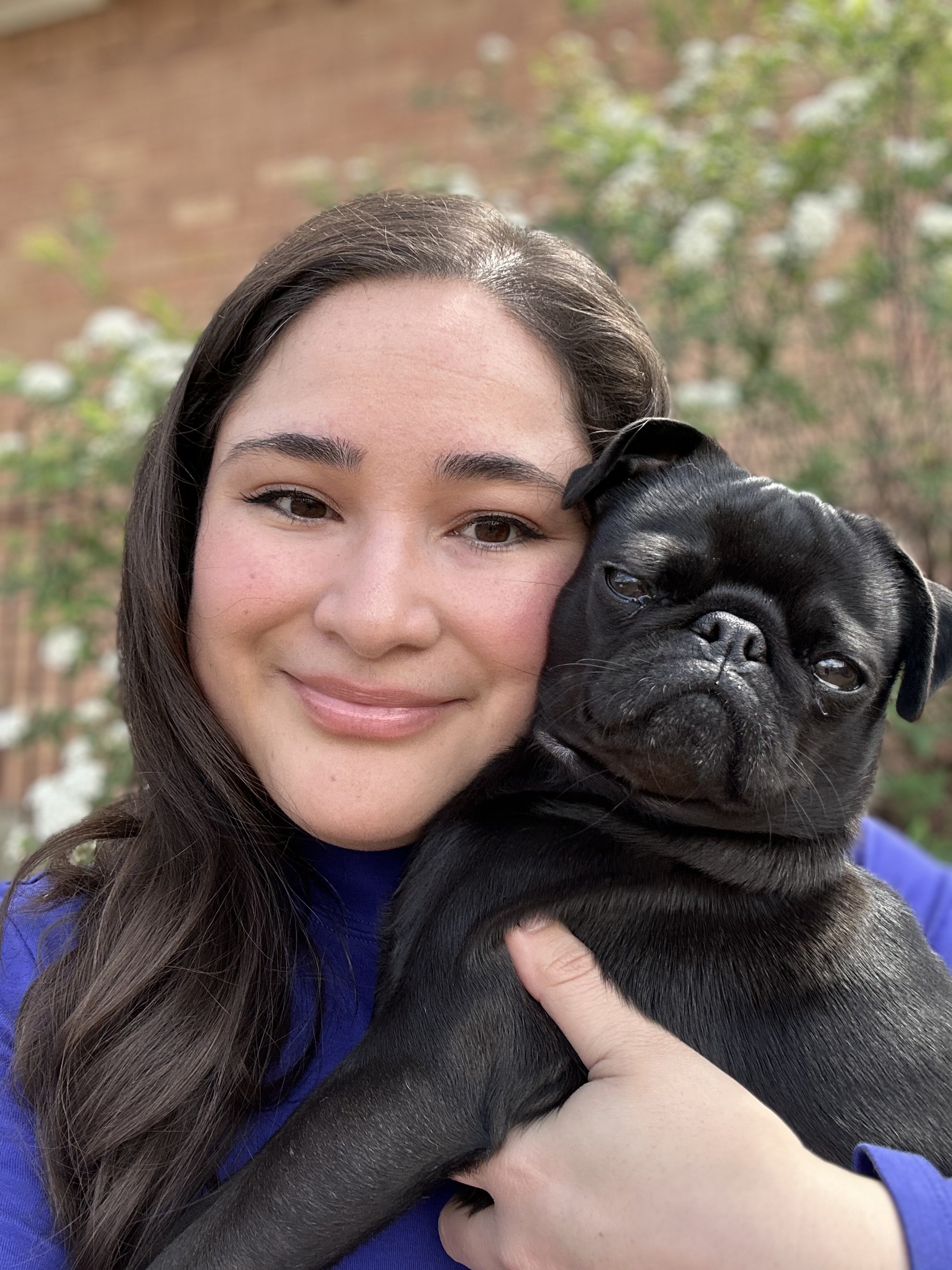 A woman smiling and holding a black pug puppy outdoors with a brick wall and greenery in the background.