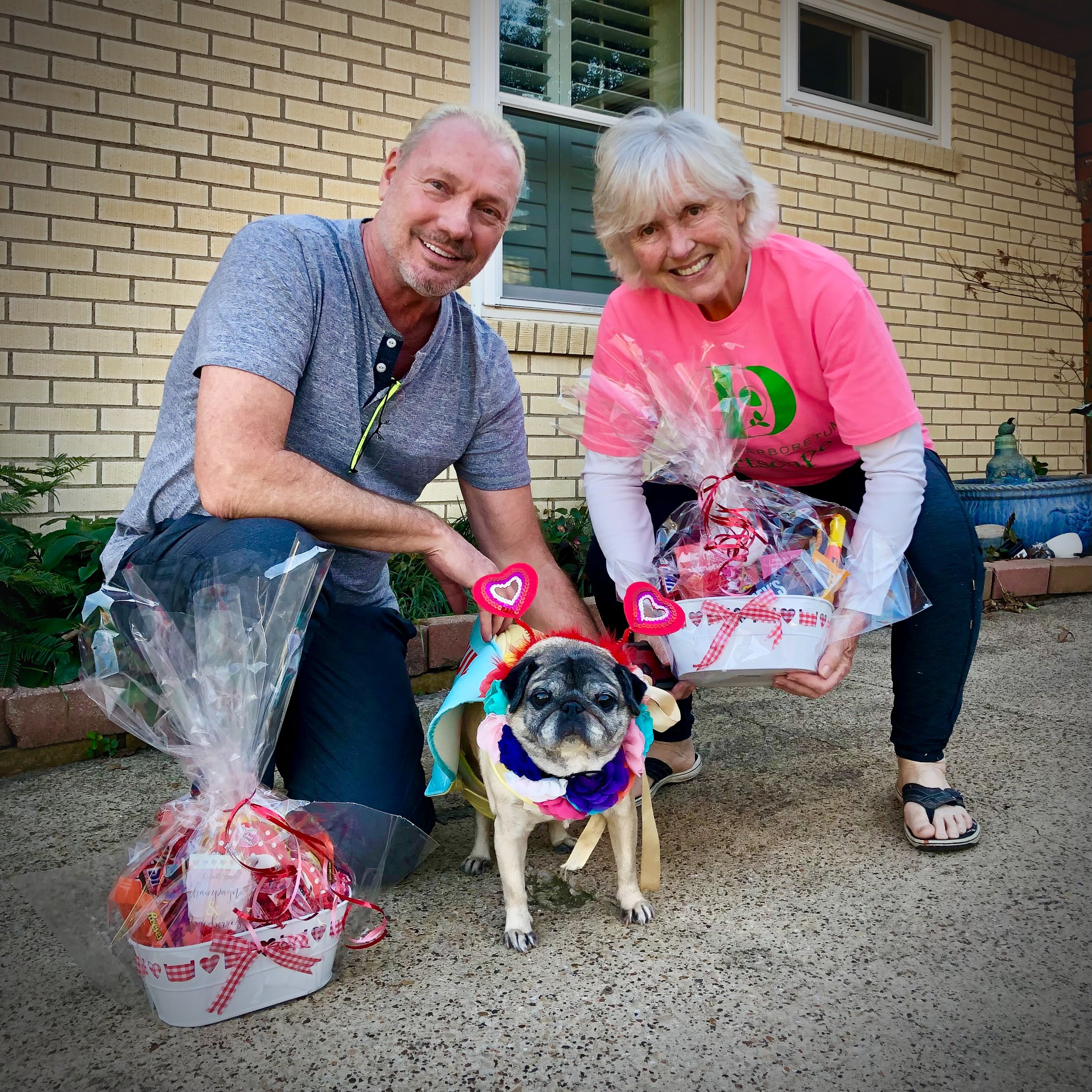 Smiling man and woman with a decorated pug dog in between, all dressed in festive attire, outdoors in front of a house, with gift baskets wrapped in cellophane and ribbon.
