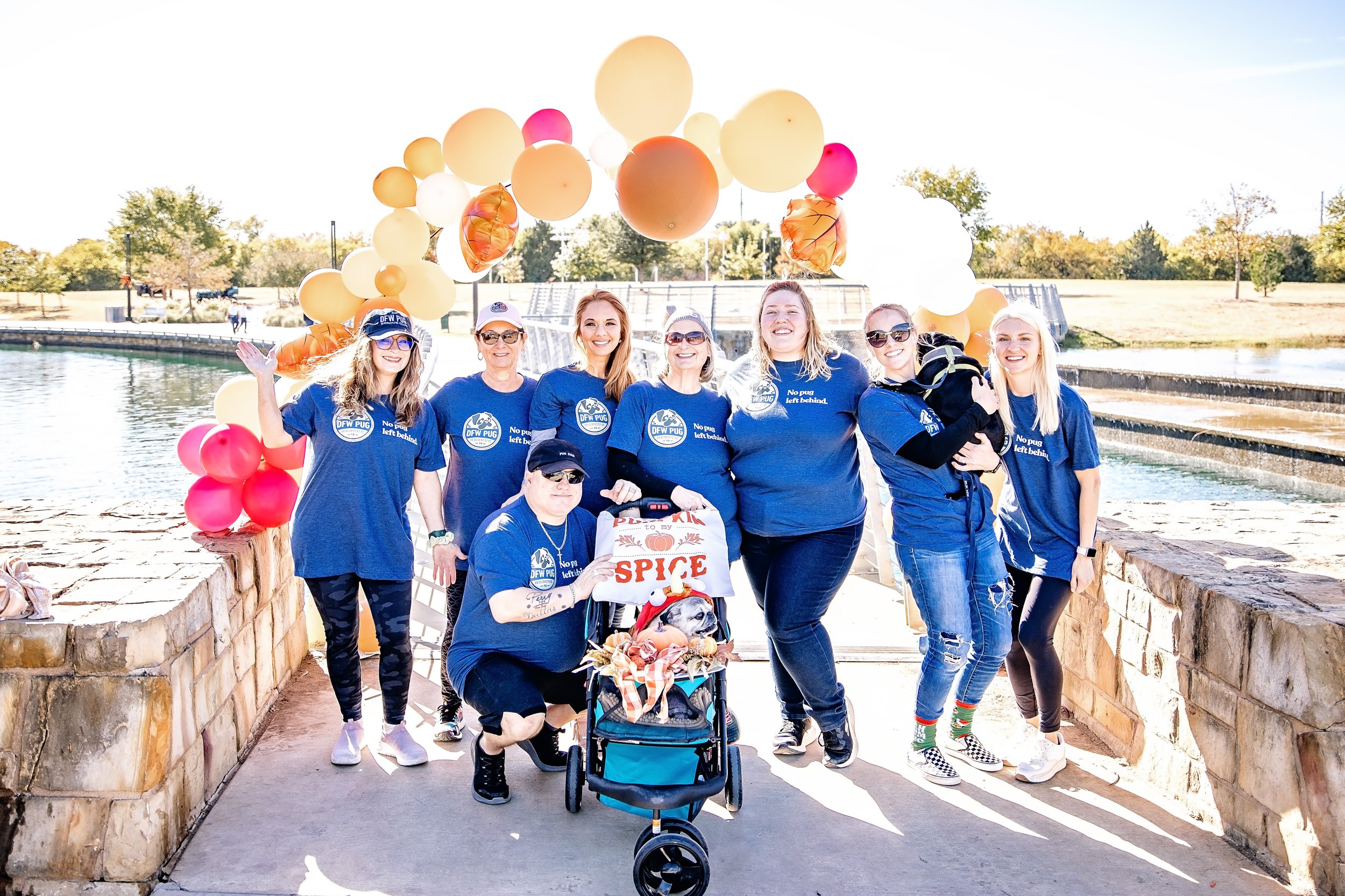 Group of people, mostly women, wearing blue shirts and sunglasses, standing outdoors in front of a water feature, celebrating with balloons and a pug dog in a stroller decorated with a sign that says "Spice." The atmosphere is cheerful, with clear sunny weather and fall foliage in the background.