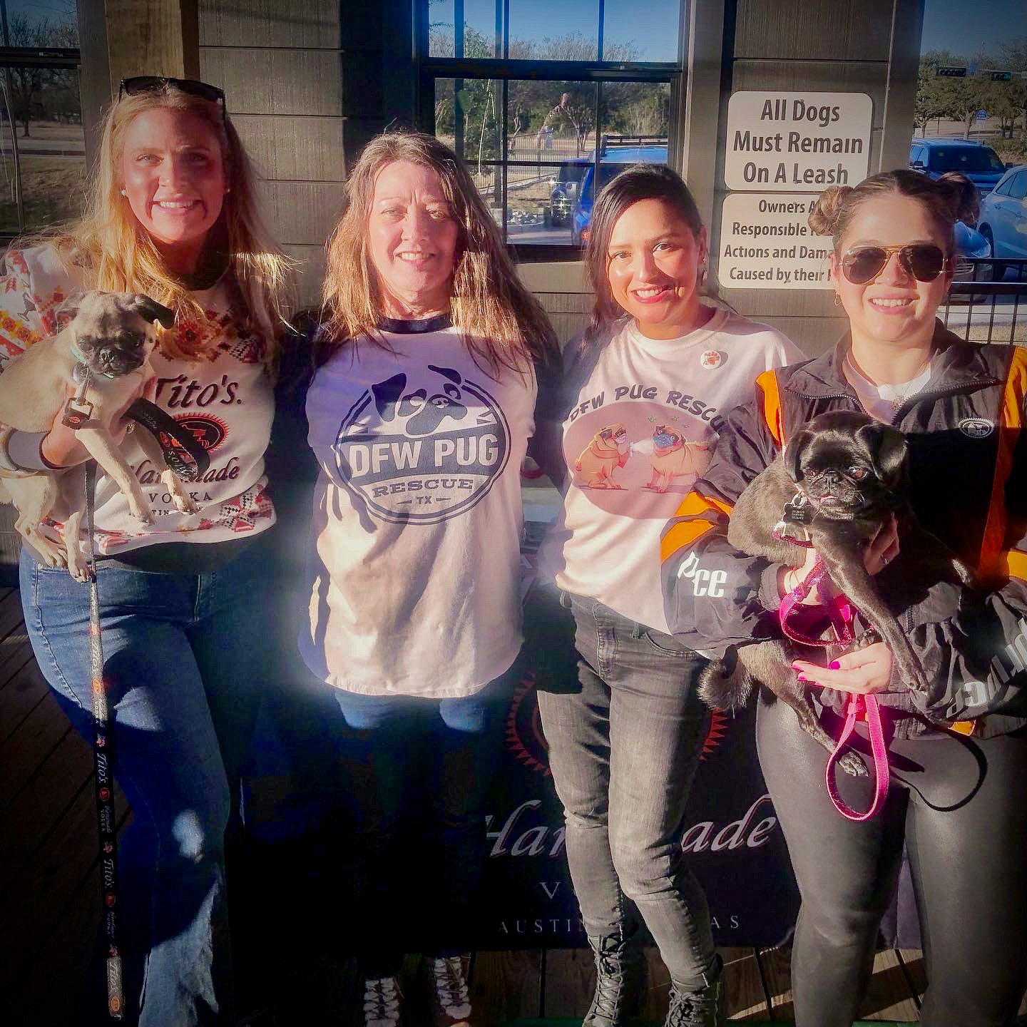 Four women standing together outdoors, holding two small dogs, with a window and a sign in the background.