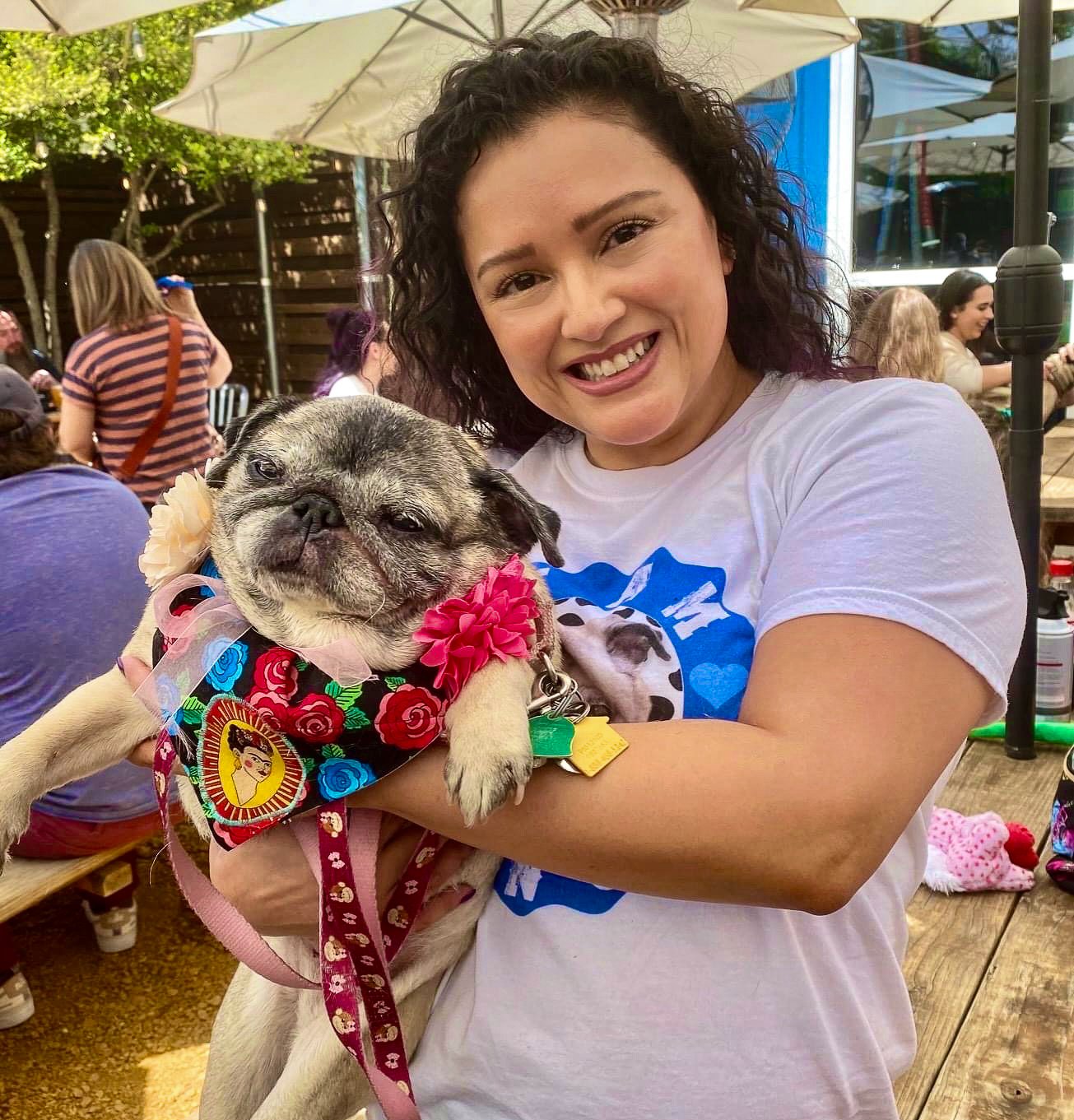 A woman smiling and holding a small, gray, wrinkly dog wearing a colorful bandana with flowers and a pink ribbon, outdoors at a social gathering.