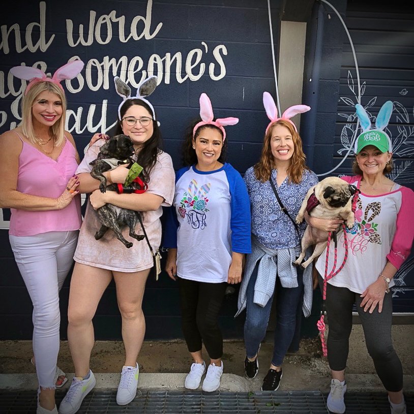 Group of five women celebrating Easter, wearing bunny ears headbands, two holding small dogs, standing in front of a wall with Easter-themed writing and drawings.