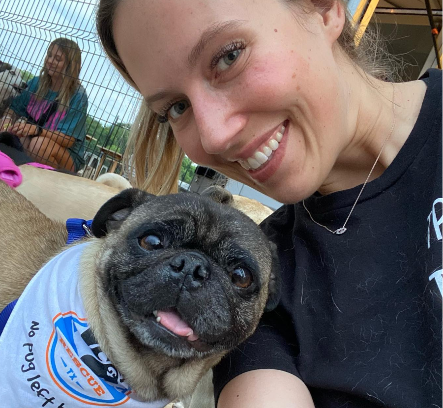A woman taking a selfie with a small pug dog. The woman is smiling, showing her teeth, and the pug has a cute expression with its tongue slightly sticking out.